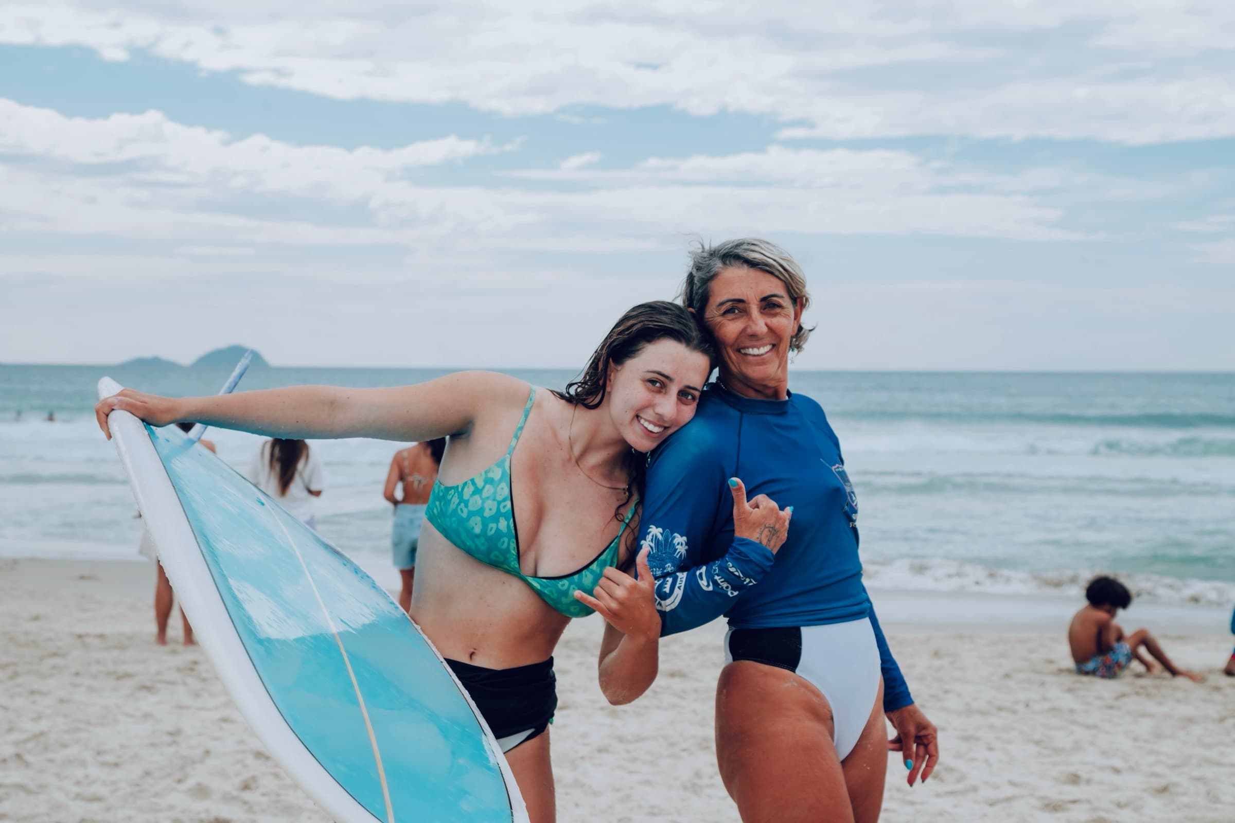 Duas mulheres sorrindo juntas na praia, uma delas segurando uma prancha.