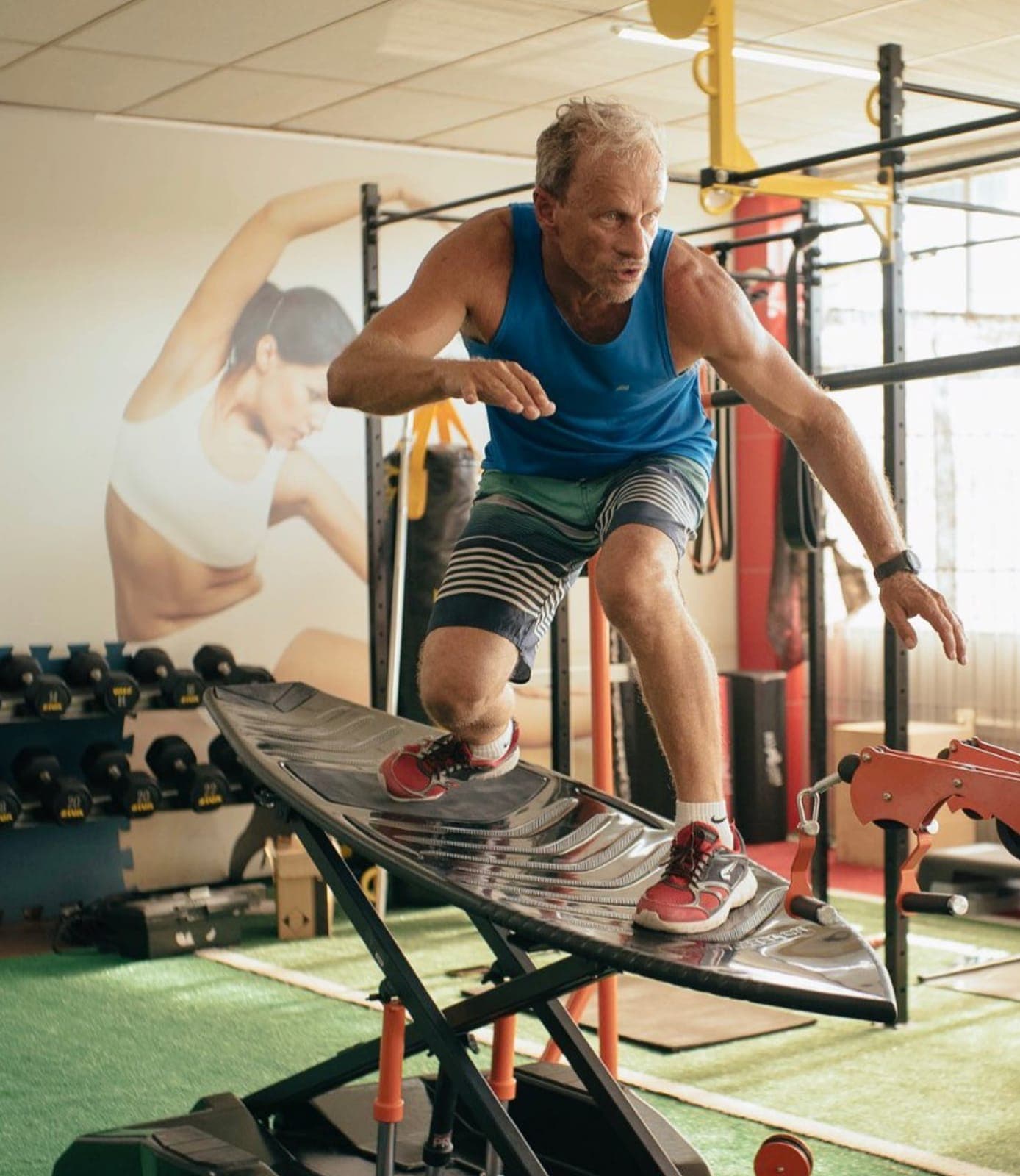Homem de regata azul se equilibrando em uma prancha de treino de surf em uma academia.