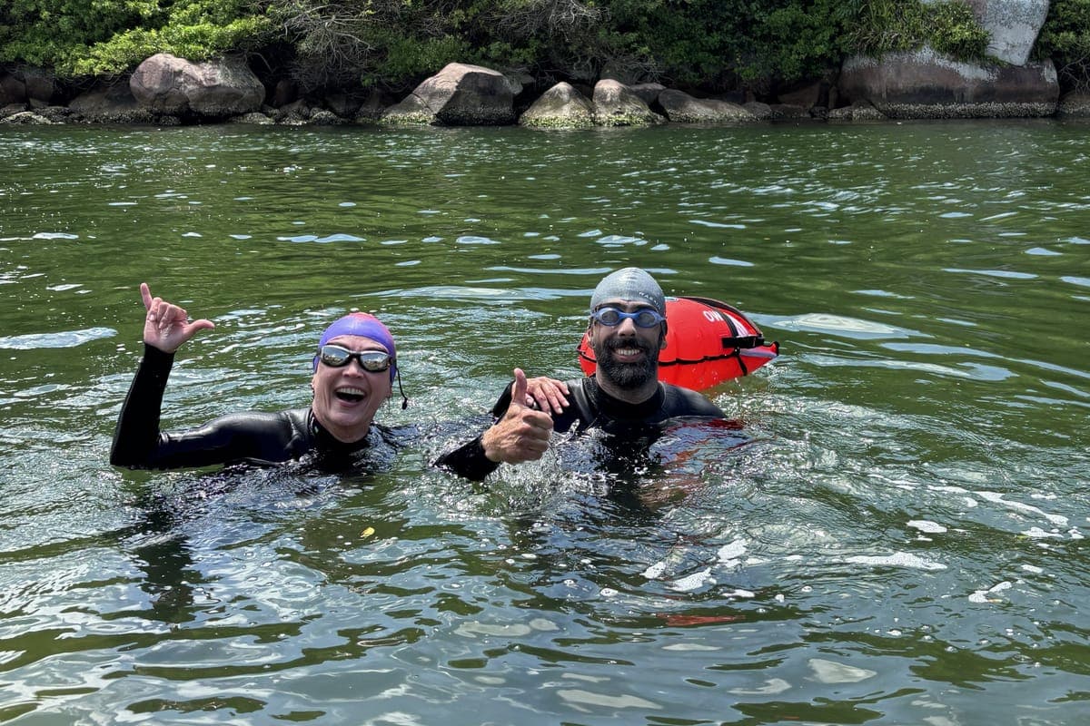 Dois nadadores de neoprene e touca em águas abertas, sorrindo com o polegar para cima, ao lado de uma boia laranja de segurança.