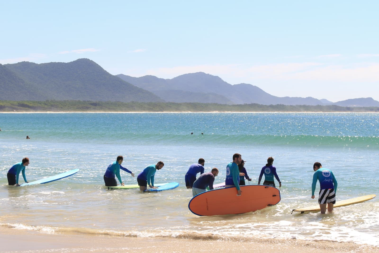 Surf Family students in blue rash guards walking into the sea with their boards during a surf lesson in Barra da Lagoa, with hills in the background.