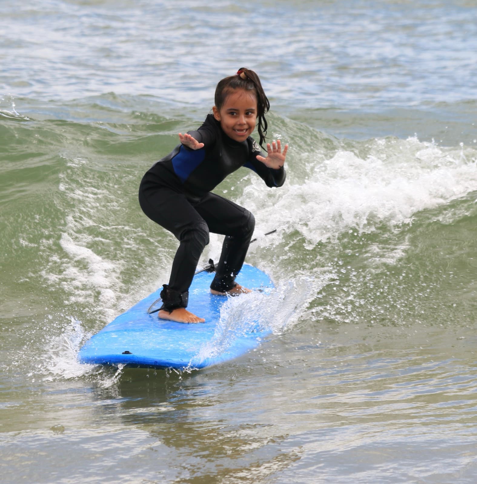 A girl in a wetsuit standing up on a small wave on a blue board in Barra da Lagoa.