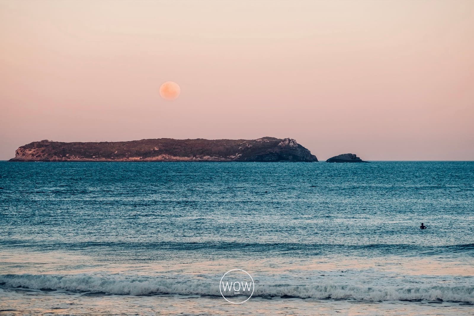 A full moon rising behind an island at dusk, seen from Joaquina beach, with one surfer waiting for a wave.