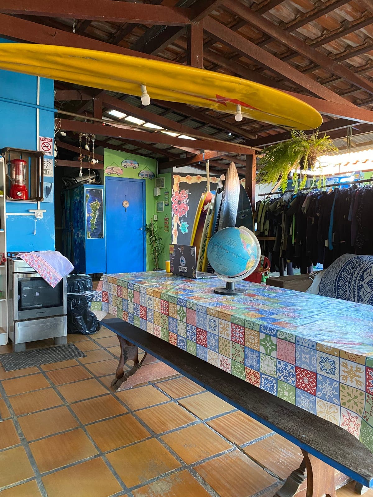 A long mosaic-tiled table with a globe, a yellow surfboard hanging overhead and wetsuits in the back.