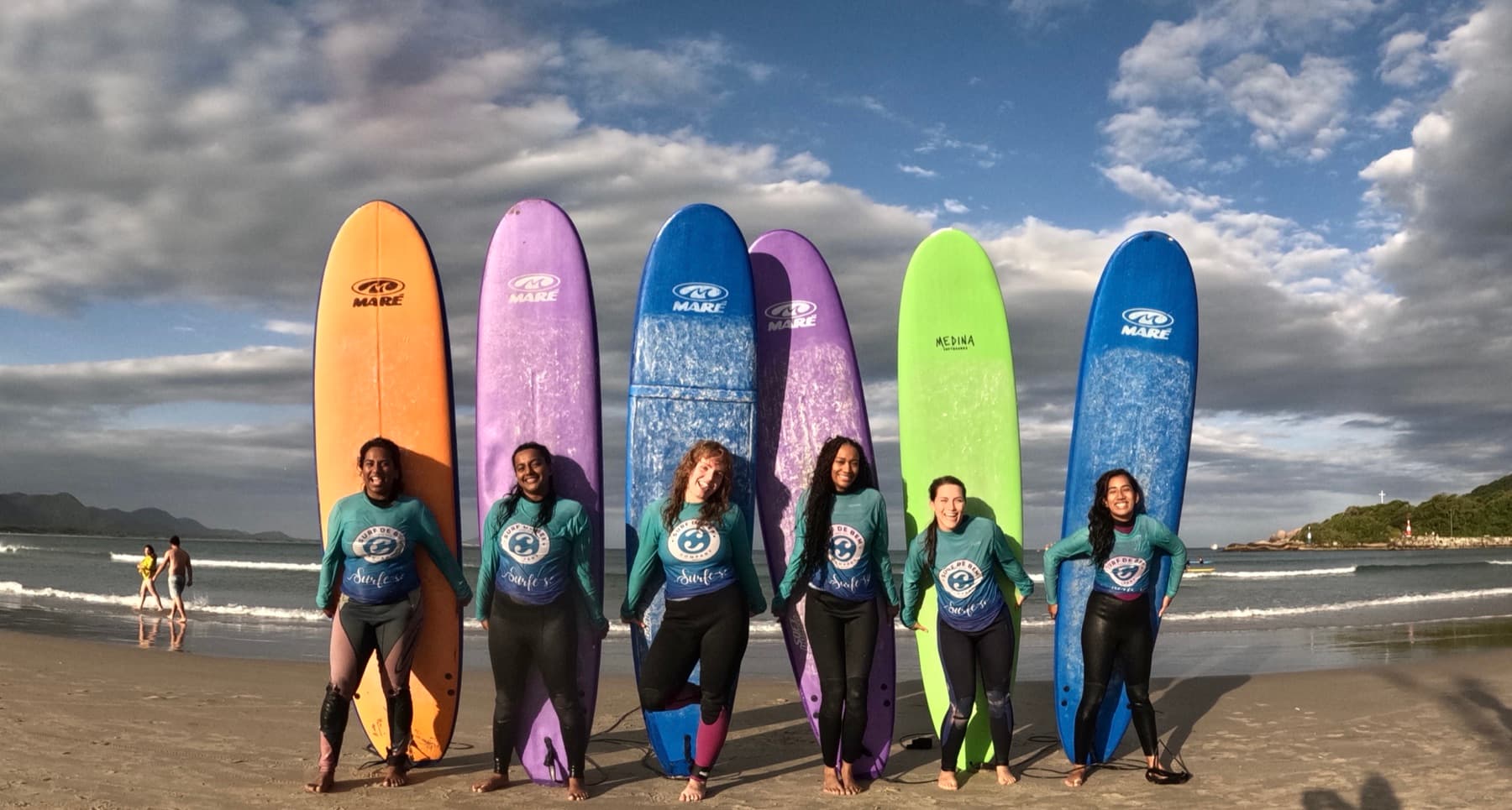 Six surfers in matching teal rash guards standing in a row in front of their upright surfboards on the beach.