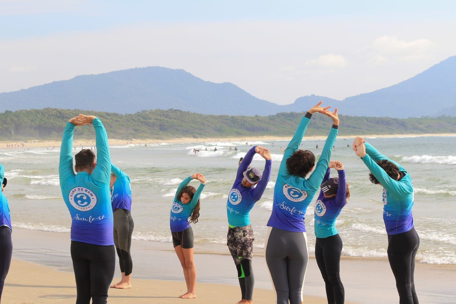 Students in matching blue rash guards stretching with their arms overhead on the beach.