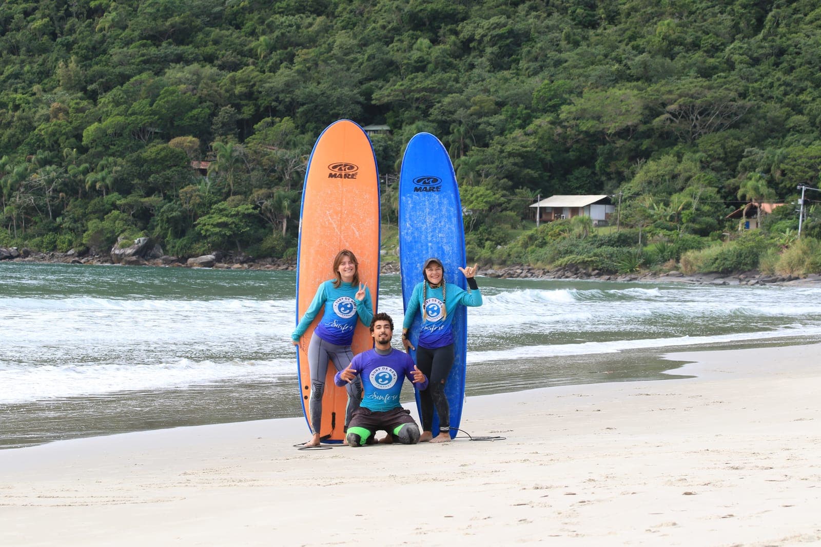 Three surfers in front of an orange and a blue board on the beach, with a green hillside behind.