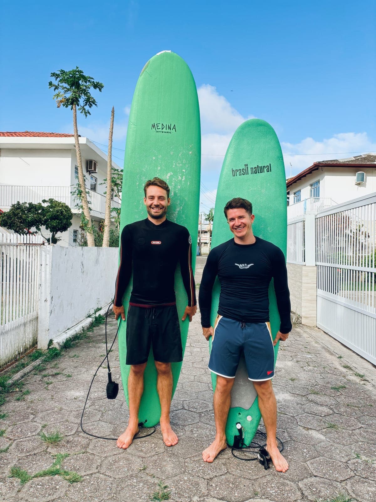 Two surfers in black wetsuits standing barefoot in front of two green surfboards.