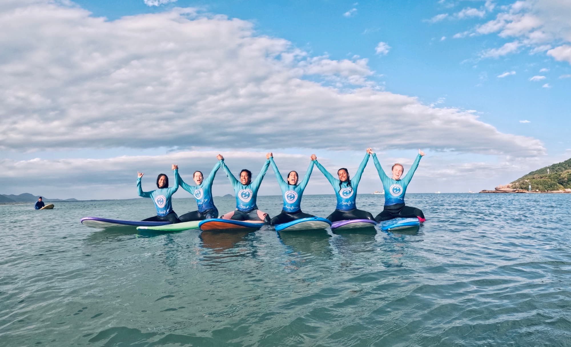Grupo de surfistas em suas pranchas na água, de mãos dadas e braços para o alto.