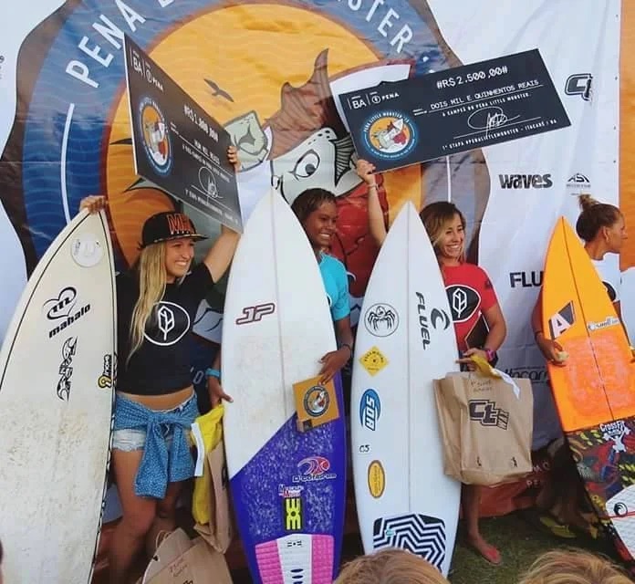 Surfistas no pódio de um campeonato com cheques de premiação e pranchas
