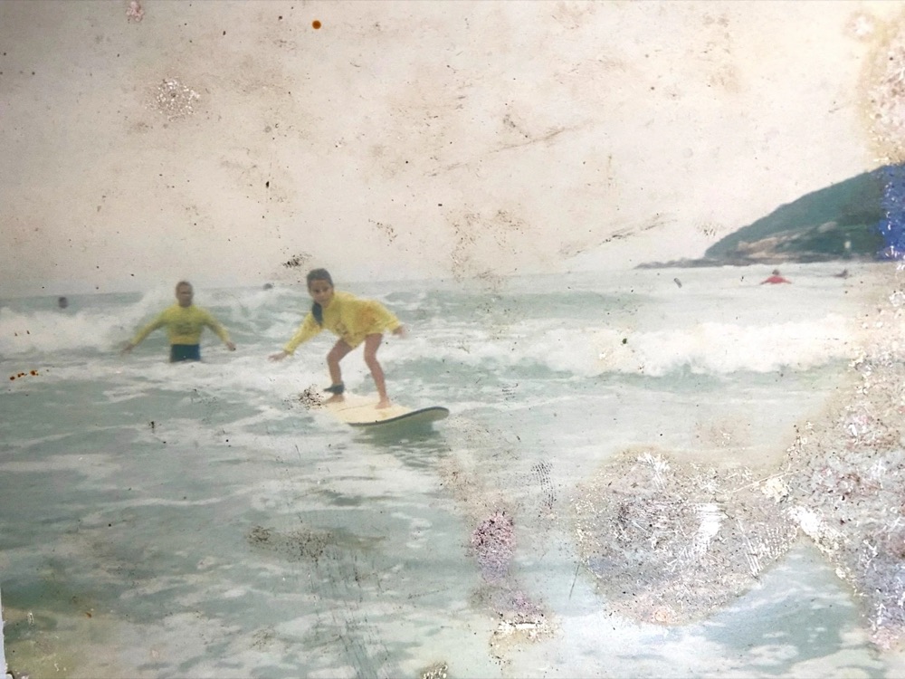 Foto antiga de uma menina surfando uma onda pequena, com um instrutor ao lado