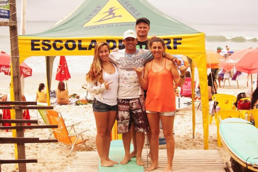 Four people posing under the school's first beach tent, with the surf school sign above them