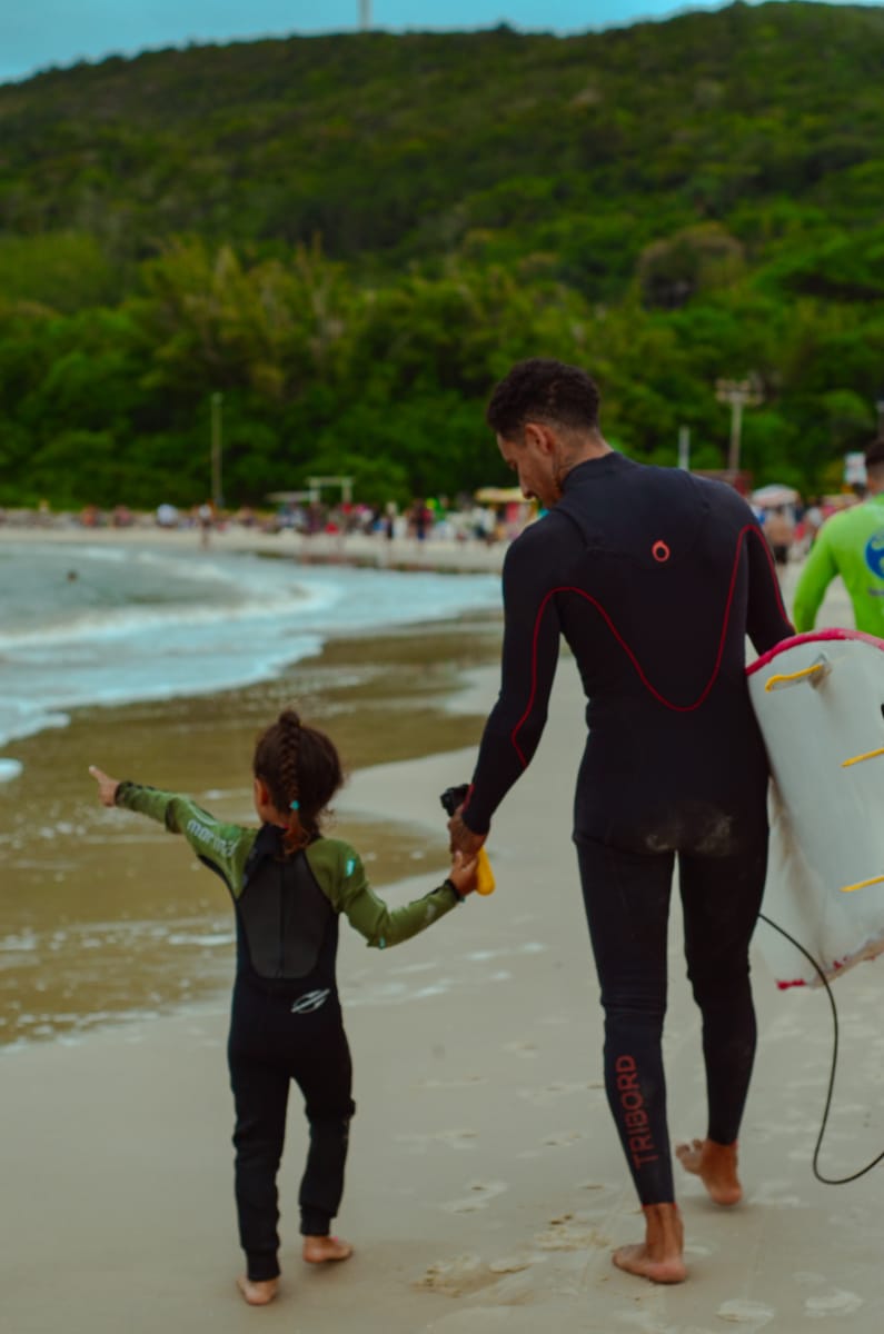 A coach in a wetsuit walking along the shore holding a young surfer's hand, both carrying boards