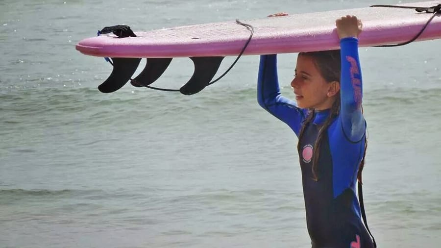 A young surfer in a blue wetsuit carrying a pink surfboard over her head at the water's edge