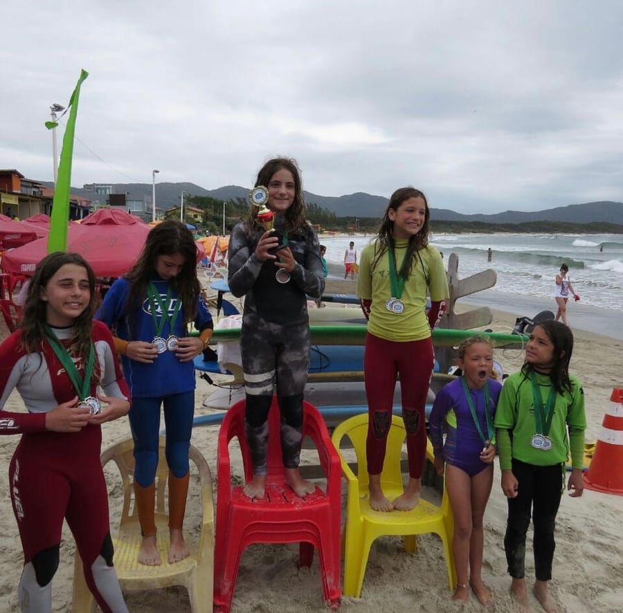 Young surfers on the beach with their medals, one standing on a chair holding a trophy