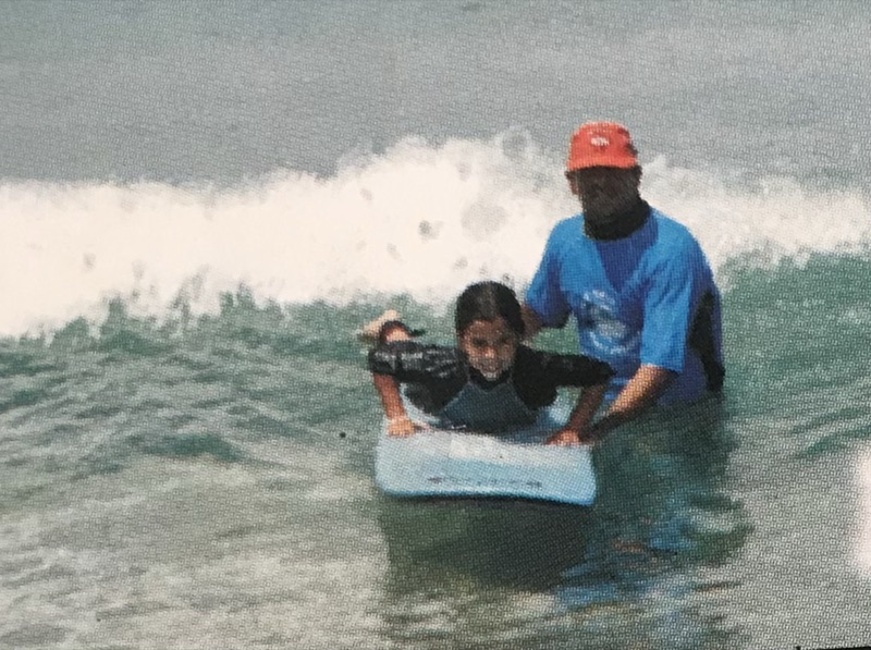 Foto antiga de uma criança deitada em uma prancha na espuma, com um instrutor em pé atrás
