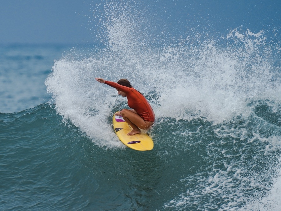 Surfista de camisa laranja fazendo uma manobra no topo da onda, em uma prancha amarela