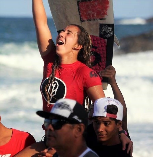 Surfista de camisa vermelha comemorando com o braço erguido, com pessoas ao redor